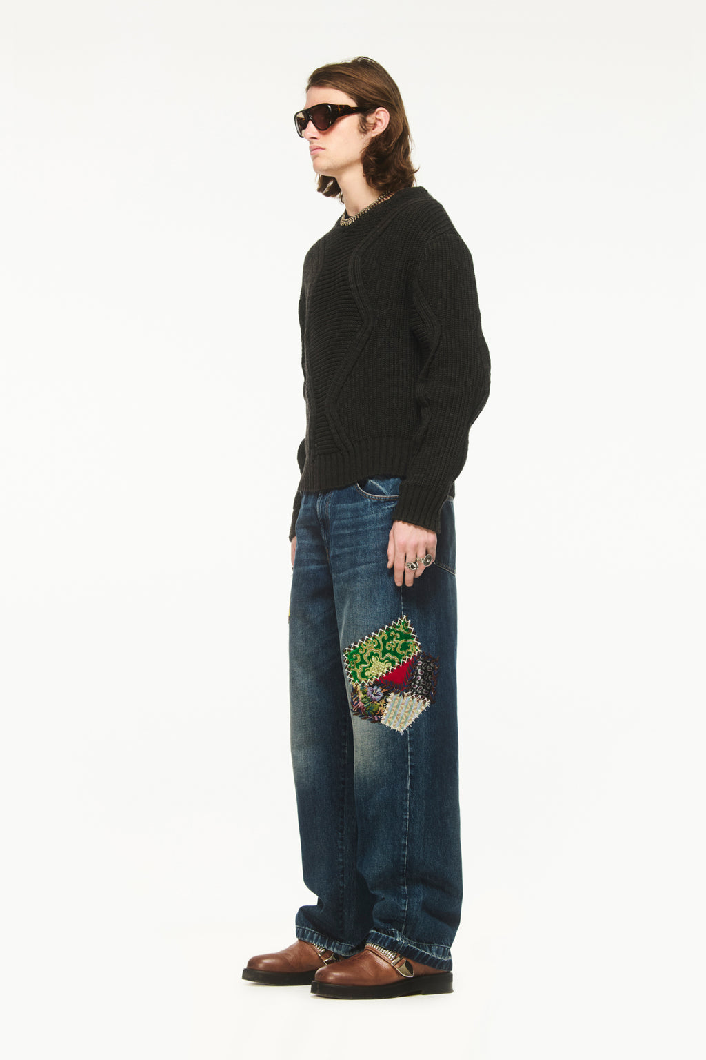 Male model wearing BLUEMARBLE dark grey chunky merino wool jumper featuring horizontal and vertical rib-knit detailing, zigzag patterns throughout the body and sleeves, ribbed crew neck, cuffs and hem, and a signature BLUEMARBLE emblem at the back styled with patchwork Bluemarble denim pants. 