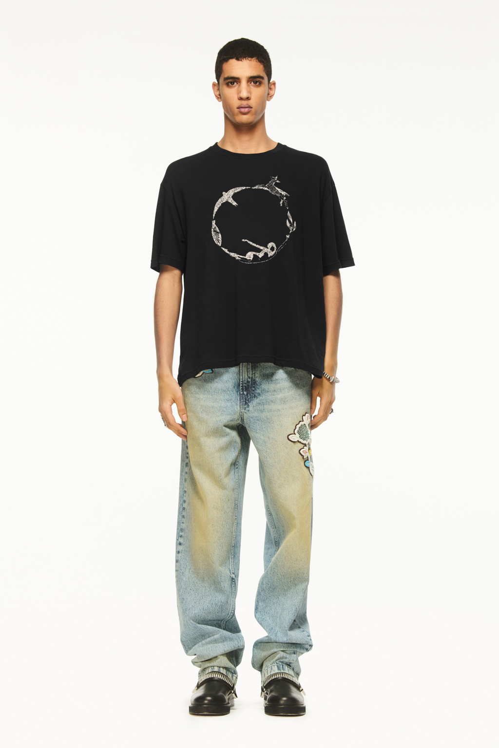 Male model wearing a BLUEMARBLE black oversized T-shirt featuring the Circle of Life graphic printed on the front, with a relaxed boxy fit and short sleeves, styled with light-wash blue jeans against a white background.