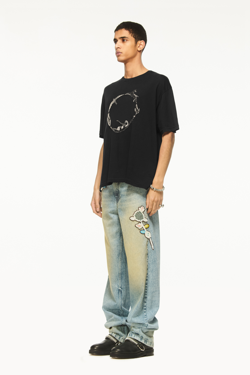 Male model wearing a BLUEMARBLE black oversized T-shirt featuring the Circle of Life graphic printed on the front, with a relaxed boxy fit and short sleeves, styled with light-wash blue jeans against a white background.