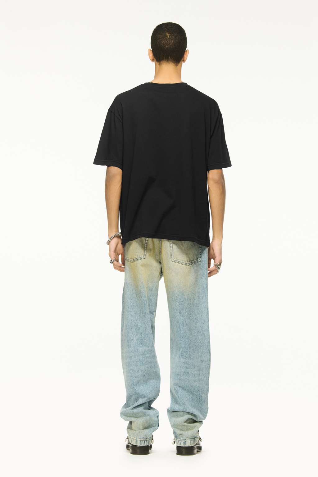 Back view of a male model wearing a BLUEMARBLE black oversized T-shirt featuring the Circle of Life graphic printed on the front, with a relaxed boxy fit and short sleeves, styled with light-wash blue jeans against a white background.