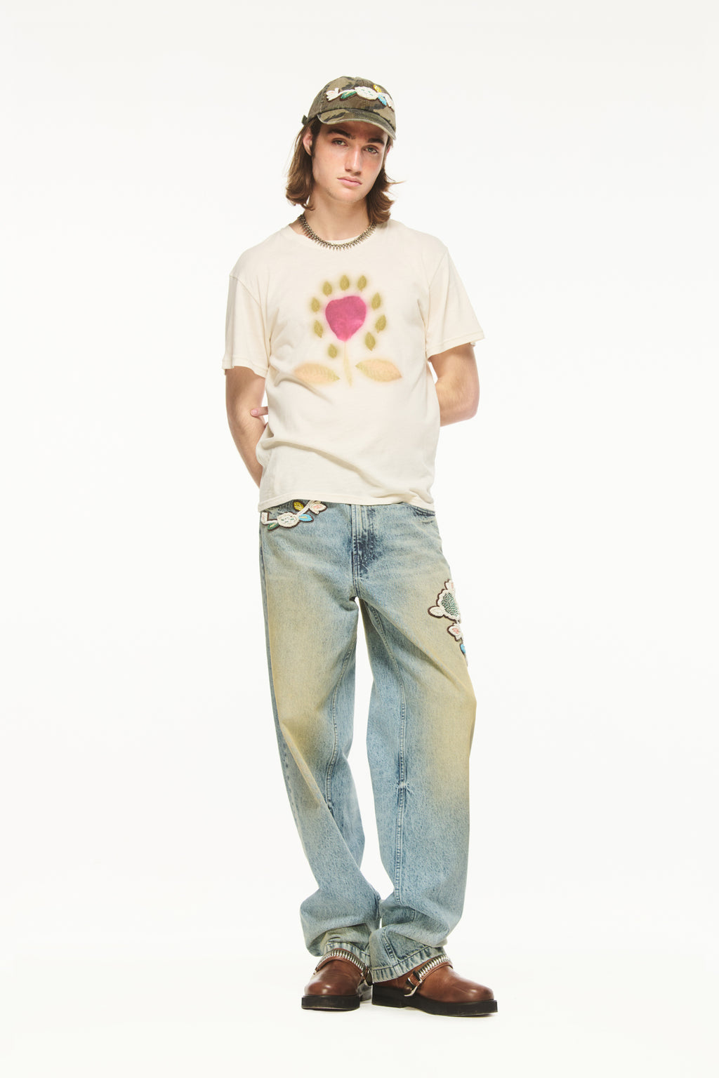 Male model with shoulder-length brown hair wearing a BLUEMARBLE washed cotton camouflage baseball cap featuring hand-embroidered multicolored botanical beading on a leather patch, styled with a spiked silver necklace and a cream T-shirt with a pink and yellow graphic against a white studio background.