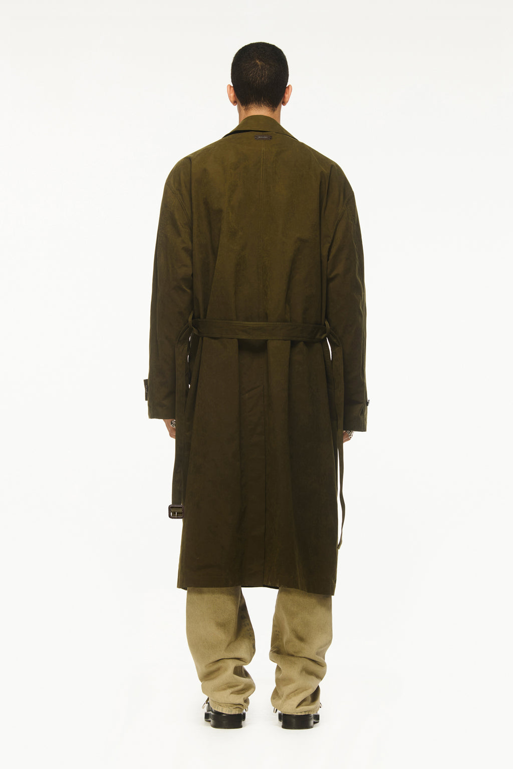 Male model wearing a BLUEMARBLE khaki water-repellent trench coat featuring a relaxed longline fit, asymmetric funnel-neck collar and belted waist with brown leather buckle, styled with a black knit sweater, beige wide-leg pants and black shoes against a white studio background.
