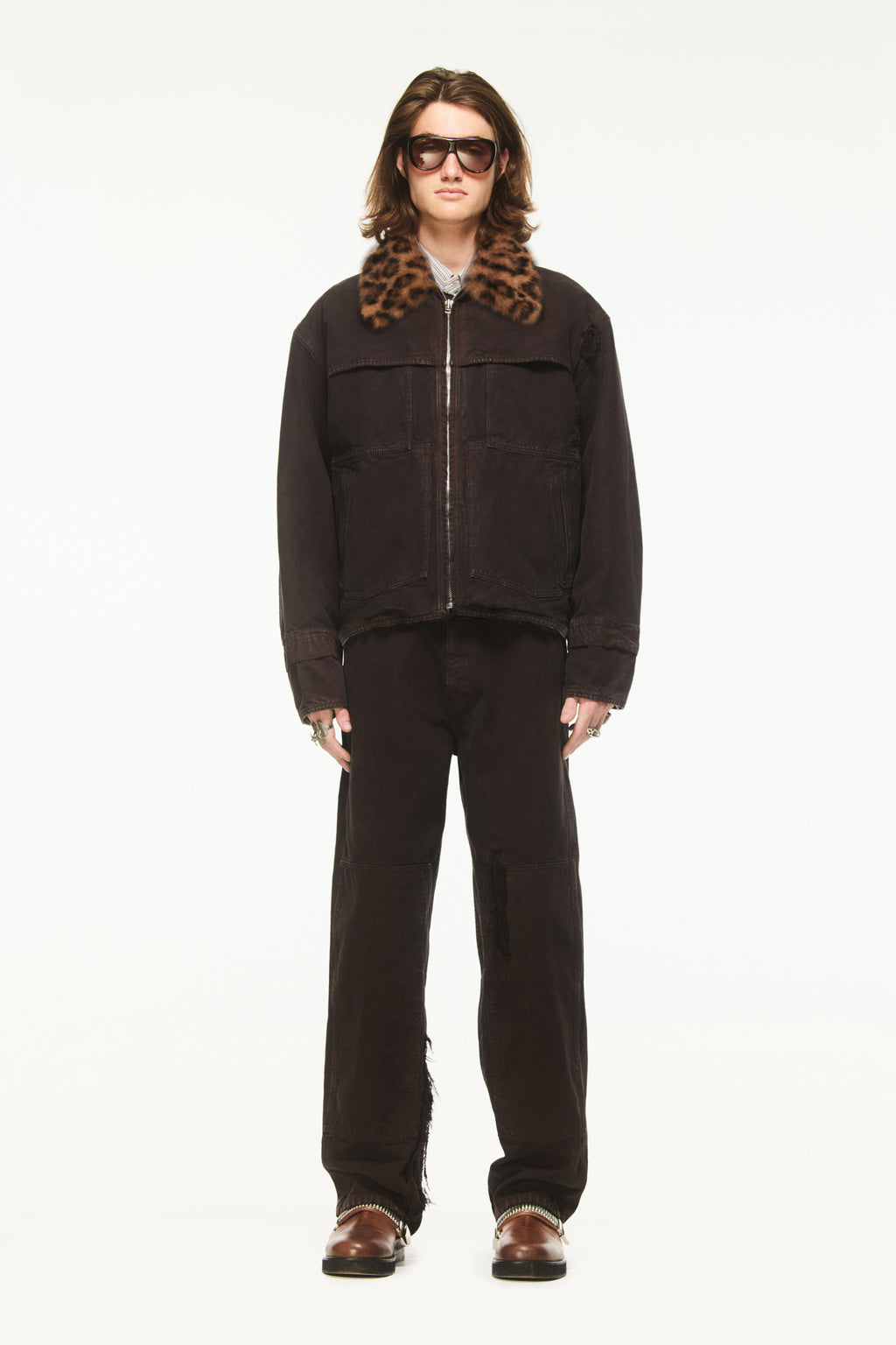 Male model wearing a BLUEMARBLE brown garment-dyed canvas workwear jacket featuring a boxy fit, detachable leopard-print faux fur collar, four front pockets and zip closure, styled with matching brown pants, a light-colored collared shirt, brown leather shoes and sunglasses against a white studio background.