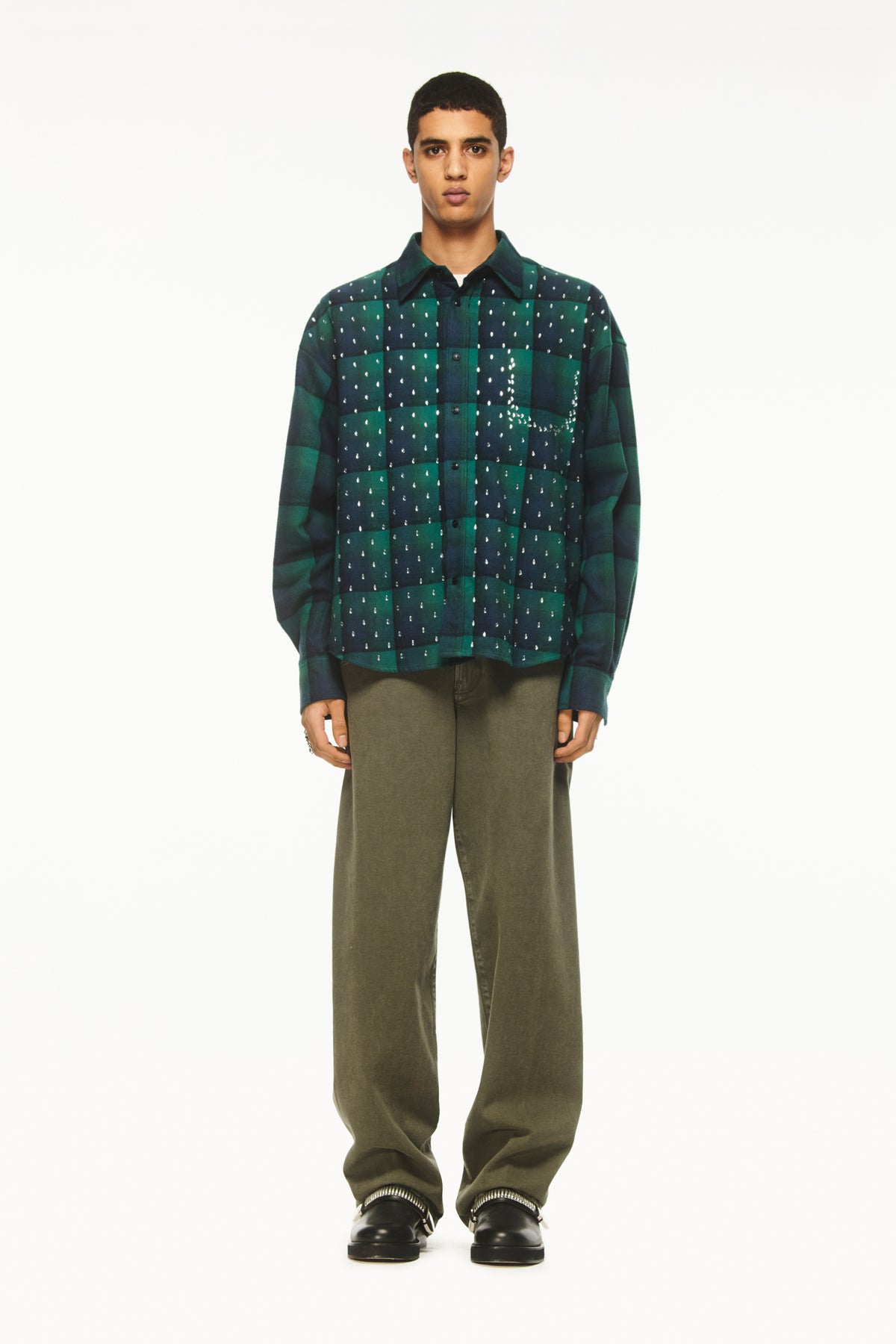 Male model wearing a BLUEMARBLE green and navy checked shirt in brushed cotton flannel, featuring rhinestone raindrop embellishments across the front, a slightly oversized fit and front button closure, styled with olive green trousers and black shoes against a white studio background.