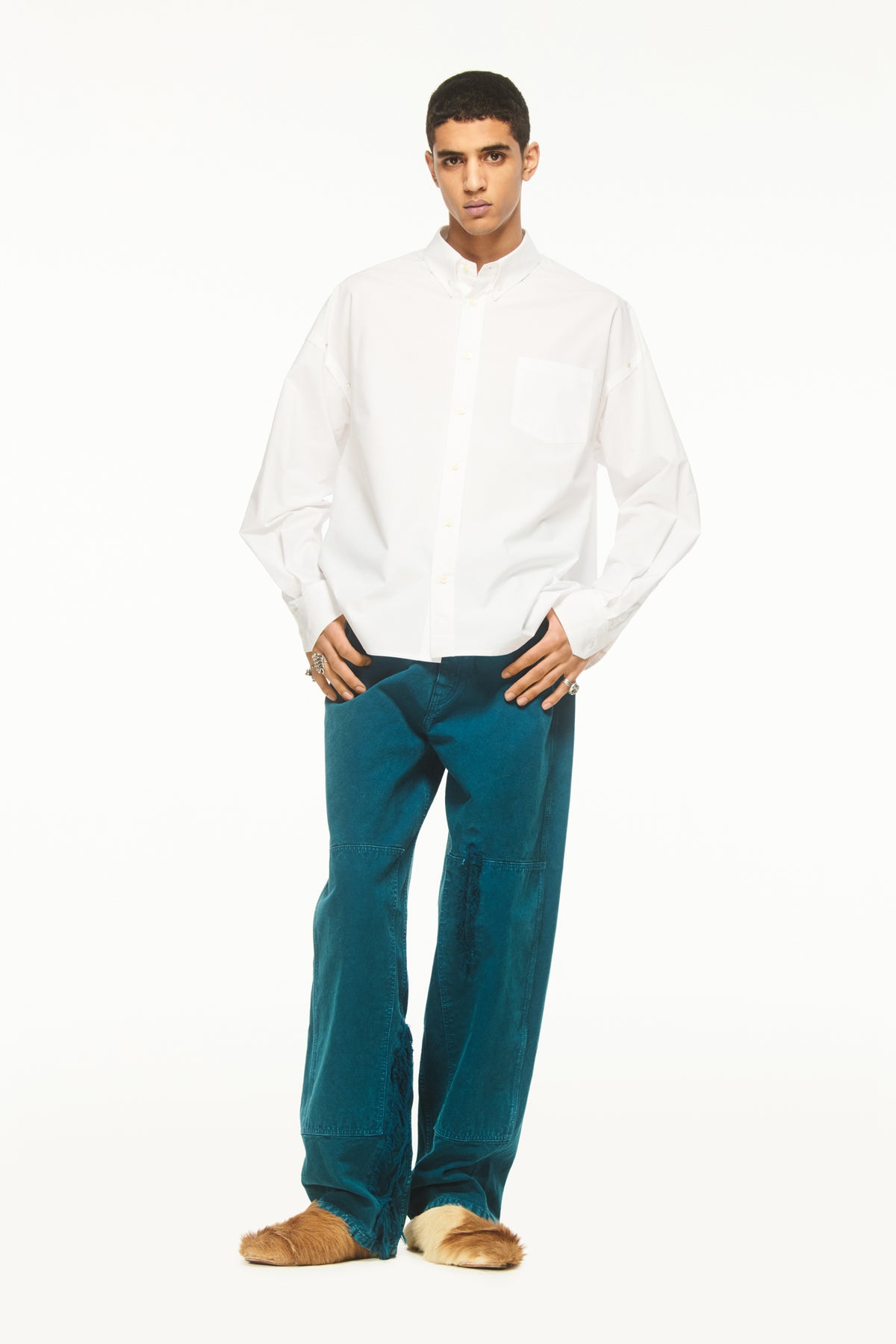Male model wearing a BLUEMARBLE white poplin shirt featuring molten metal studs along the shoulders and armholes, raw-edge detailing and a slightly oversized fit, styled with duck green workwear pants against a white studio background.