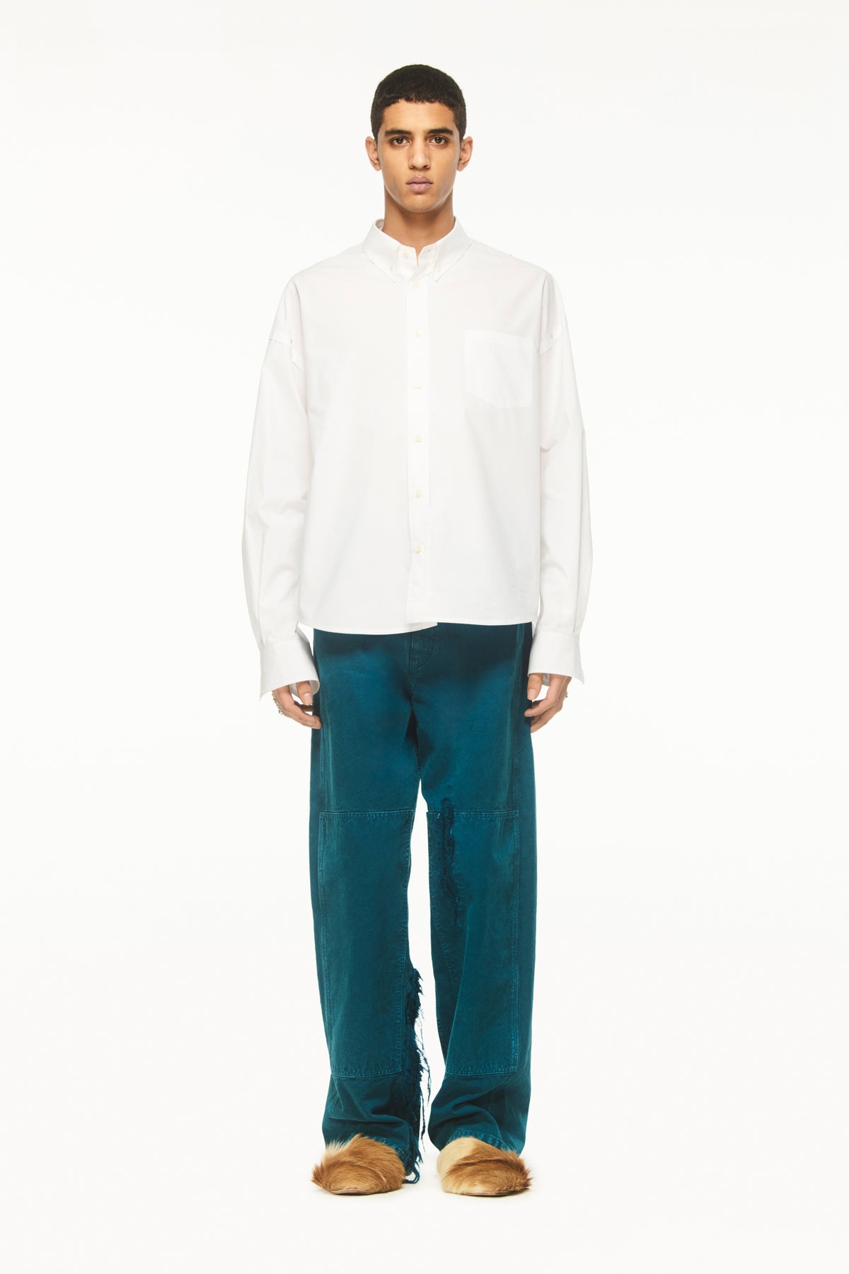 Male model wearing a BLUEMARBLE white poplin shirt featuring molten metal studs along the shoulders and armholes, raw-edge detailing and a slightly oversized fit, styled with duck green workwear pants against a white studio background.