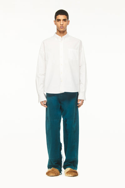 Male model wearing a BLUEMARBLE white poplin shirt featuring molten metal studs along the shoulders and armholes, raw-edge detailing and a slightly oversized fit, styled with duck green workwear pants against a white studio background.