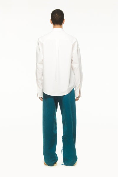 Male model wearing a BLUEMARBLE white poplin shirt featuring molten metal studs along the shoulders and armholes, raw-edge detailing and a slightly oversized fit, styled with duck green workwear pants against a white studio background.
